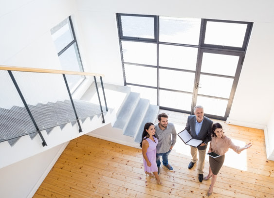four people standing in a bright, empty room with large windows and a staircase, looking up toward the camera