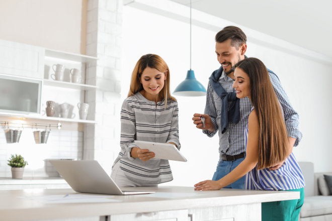 three people gathered around a kitchen counter, smiling while looking at a tablet